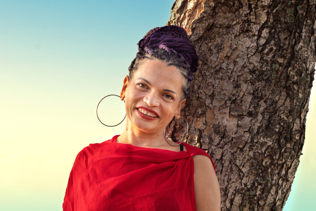 Retrato de Jeannette sonriente con cabello trenzado y vestido rojo junto a un árbol, bajo cielo azul y dorado al atardecer