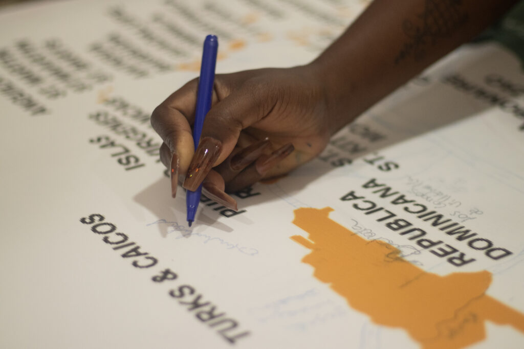 Mano escribiendo con bolígrafo azul sobre mapa con nombres de países del Caribe, incluyendo República Dominicana y Turks & Caicos.