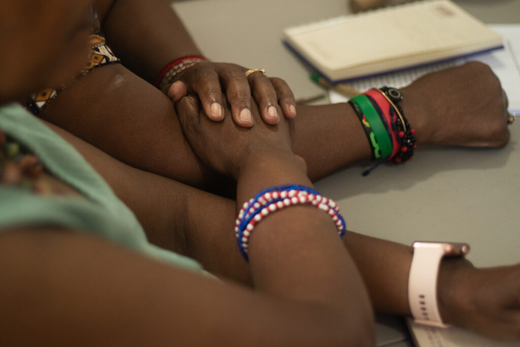Dos personas afrodescendientes con pulseras coloridas y reloj inteligente descansan sus manos sobre una mesa junto a una libreta, en un ambiente de diálogo y colaboración.